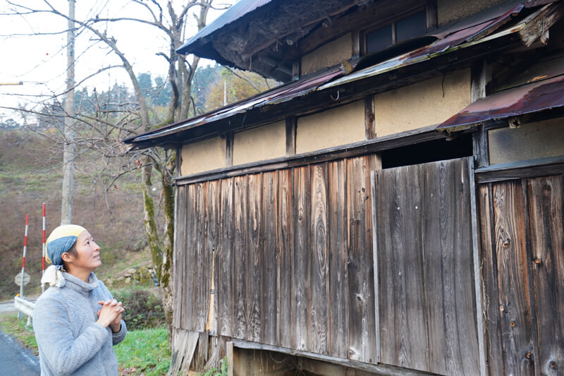 藁葺きの屋根にトタンをかぶせた古い家屋の前で説明する小山さん